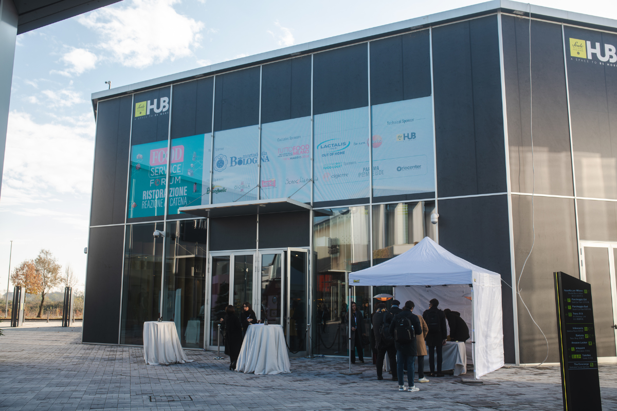 Ingresso al Food Service Forum: evento sulla ristorazione italiana. Persone in fila, stand e loghi di sponsor.