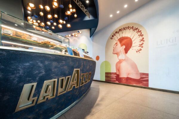 Design Lavazza Coffee all'aeroporto di Verona. Bancone bar blu con logo Lavazza dorato, murales artistico e illuminazione moderna.