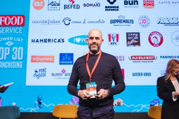 Premiazione Food Service Powerlist: top 30 franchising. Uomo con premio, loghi ristorazione sullo sfondo.