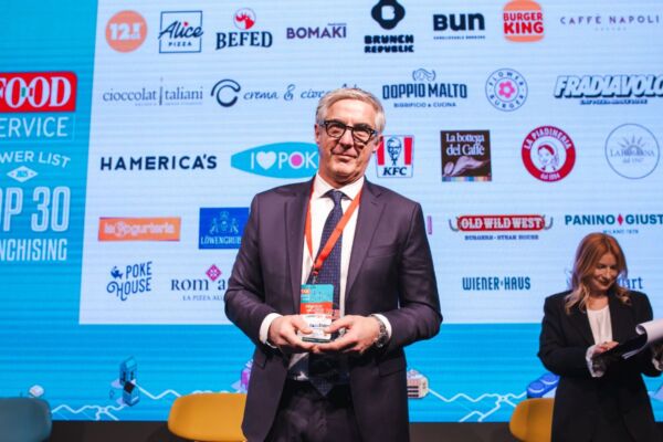 Top 30 Franchising: uomo in giacca davanti a loghi food service. Evento franchising, premiazione aziende ristorazione.