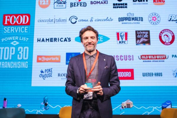 Premiazione Top 30 Franchising Food Service. Uomo con premio davanti a loghi di aziende ristorazione.