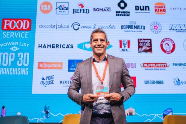 Premiazione Top 30 Franchising Food Service. Uomo sorridente con premio, marchi ristorazione sullo sfondo.