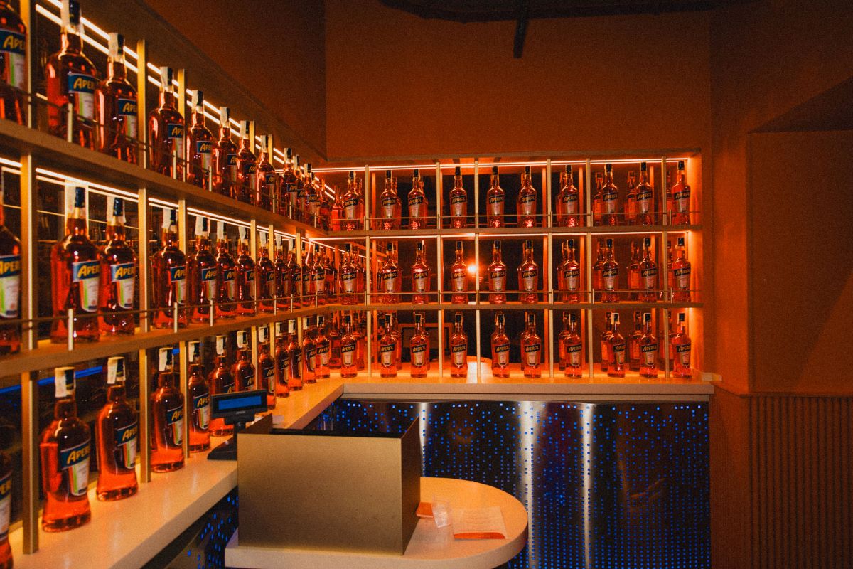 Bar SYNC by Aperol a Milano: scaffali pieni di bottiglie Aperol. Design moderno e illuminazione arancione.