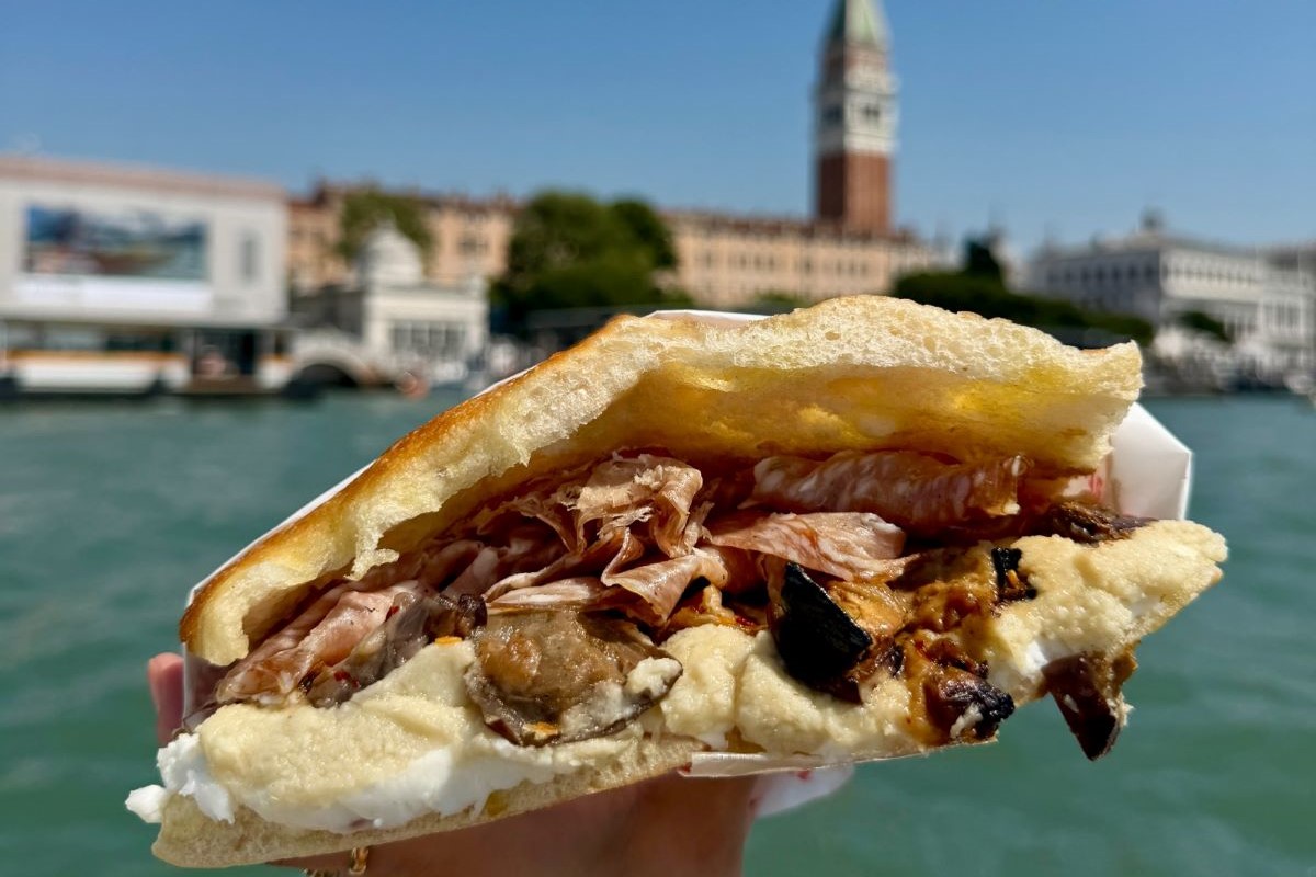 Schiacciata farcita di All'Antico Vinaio tenuta in mano davanti al panorama di Venezia con il campanile di San Marco.