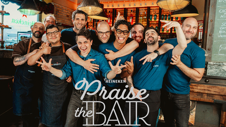 Gruppo di baristi sorridenti in un locale Heineken per la campagna Praise the Bar, atmosfera conviviale e professionale.