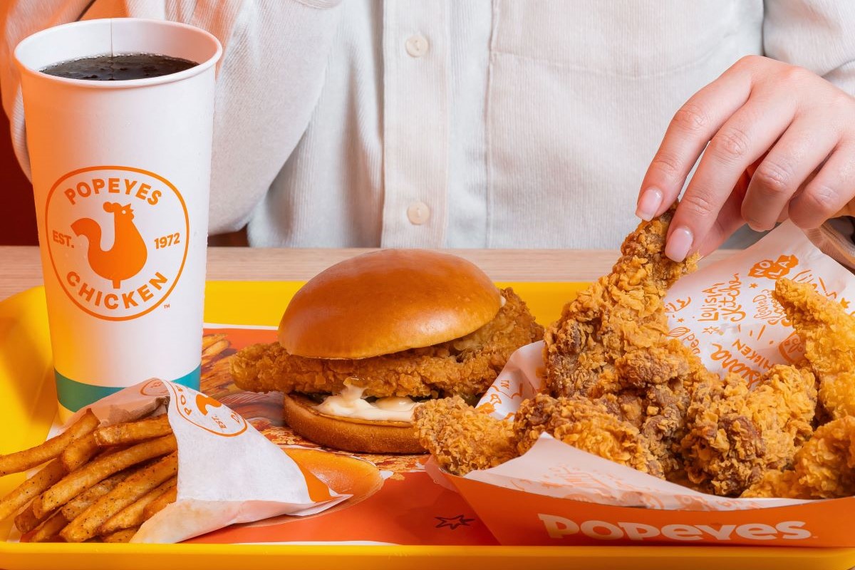 Menu Popeyes in Italia: panino con pollo fritto, patatine e bibita su vassoio giallo.