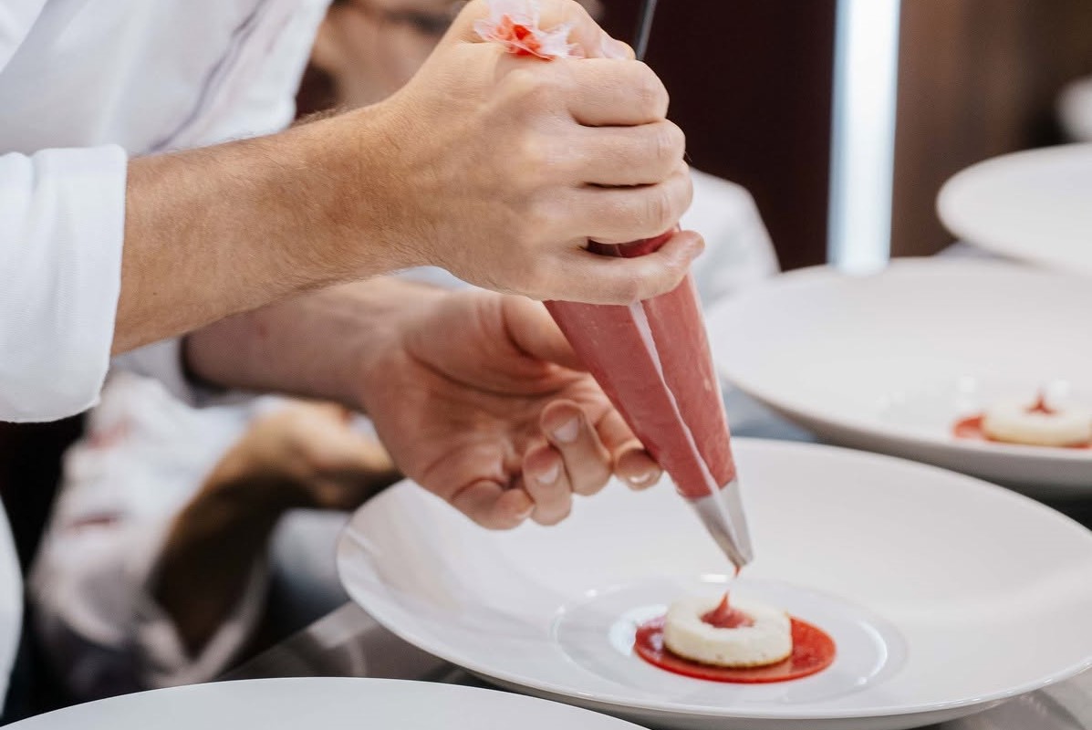 Chef pasticciere utilizza una sac-à-poche per decorare un dessert Irca su un piatto bianco in una cucina professionale.