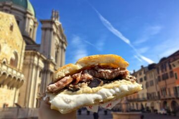 Schiacciata farcita di All'Antico Vinaio tenuta in mano davanti al Duomo di Brescia in una giornata di sole.