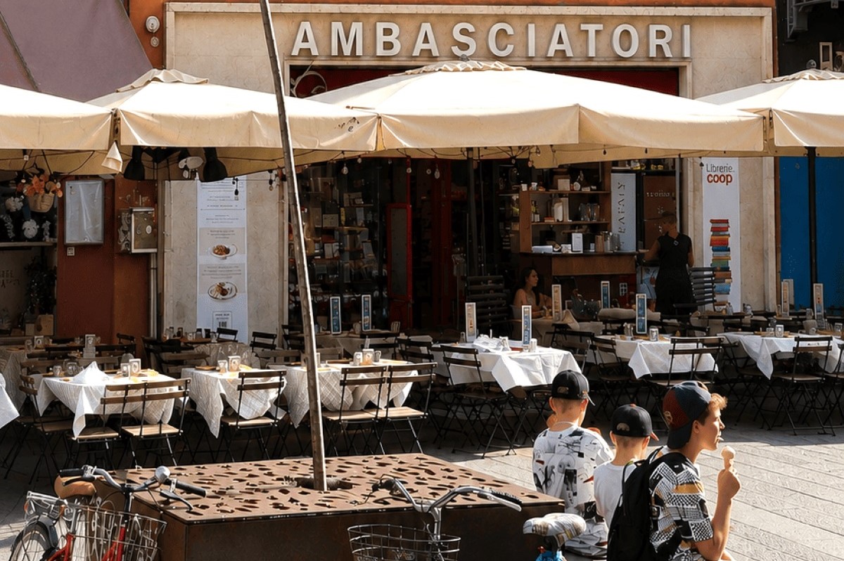 Esterno della libreria Ambasciatori Coop Alleanza 3.0 con tavolini, ombrelloni e clienti in una piazza cittadina.