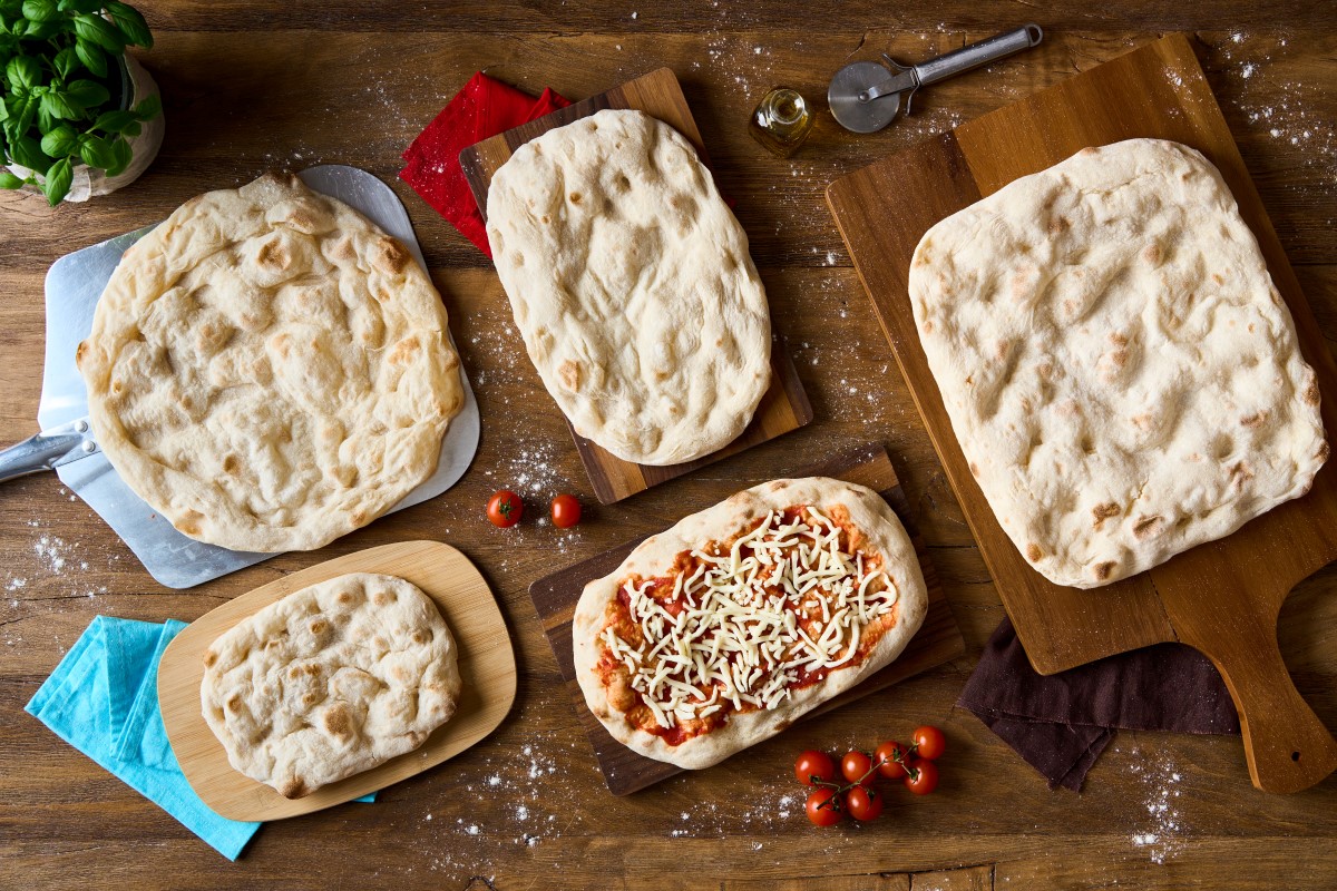 Selezione di basi per pinsa romana artigianale su taglieri di legno, con una pinsa condita, pomodorini e basilico fresco.