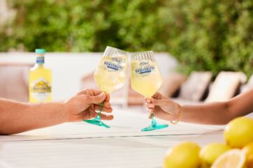 Due persone brindano con calici di Limoncello Benvenuti ghiacciato, con bottiglia e limoni freschi su un tavolo all'aperto.