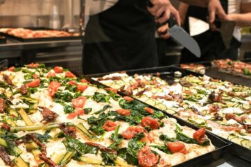 Pizzaiolo taglia teglie di pizza alla romana con verdure, pomodori e formaggio nel bar caffetteria Rom’antica.