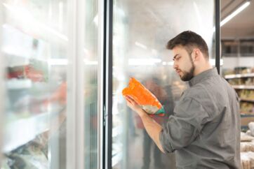 Uomo sceglie alimenti surgelati leggendo l'etichetta di una confezione davanti al banco frigo di un supermercato.