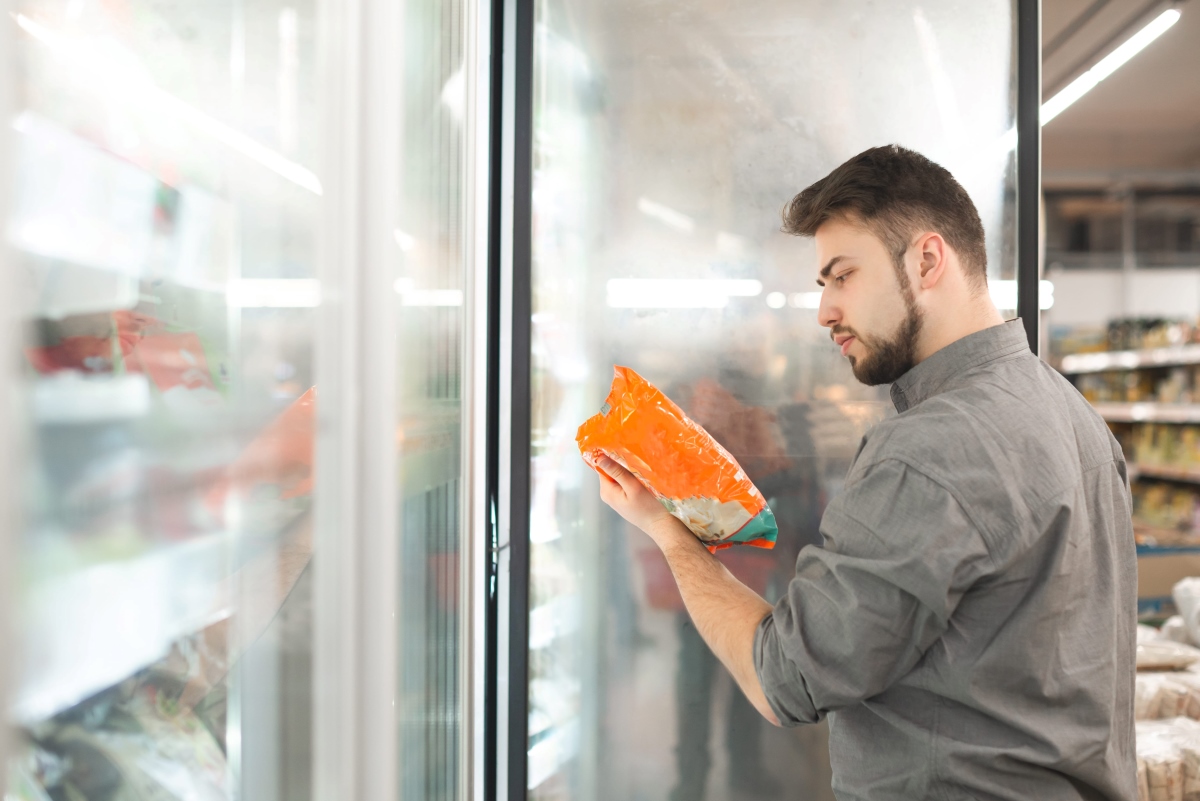 Uomo sceglie alimenti surgelati leggendo l'etichetta di una confezione davanti al banco frigo di un supermercato.