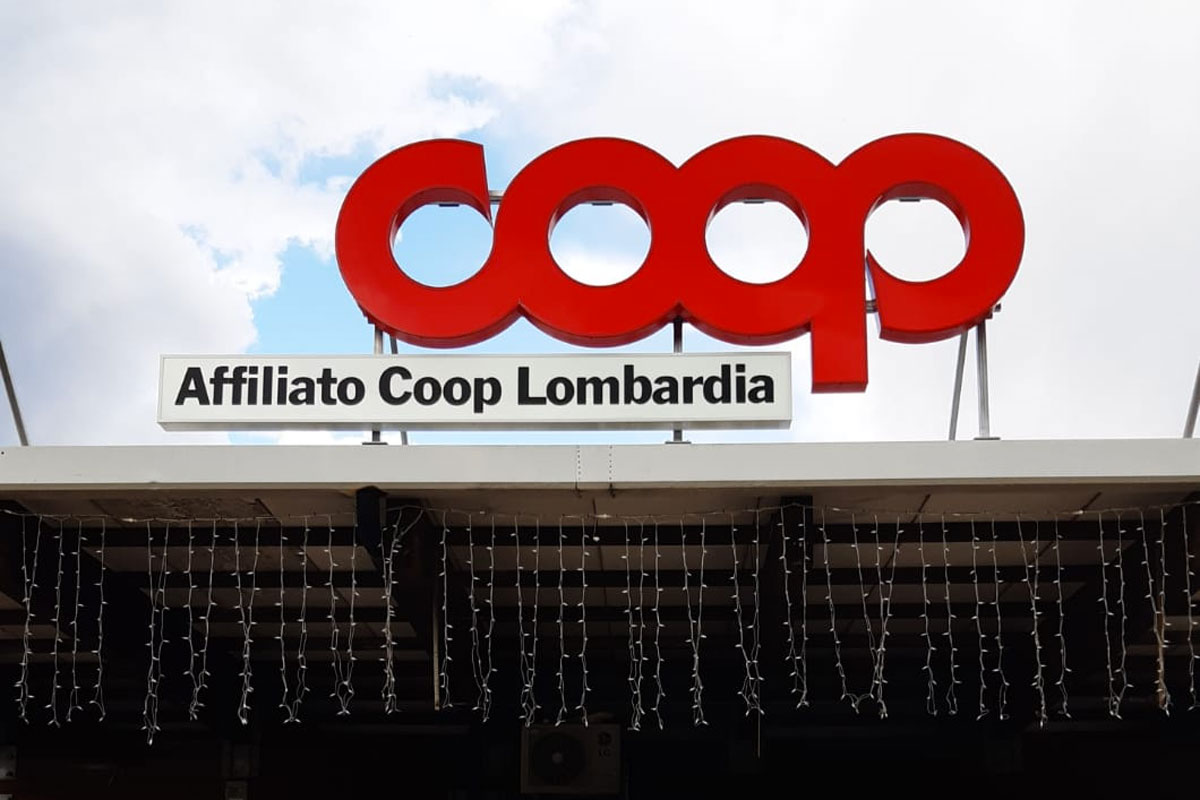 Insegna rossa Coop e scritta Affiliato Coop Lombardia sul tetto di un supermercato, con luci appese.