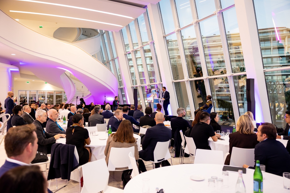 Conferenza MAPIC 2026: relatori e pubblico in sala con grandi vetrate e luci viola.
