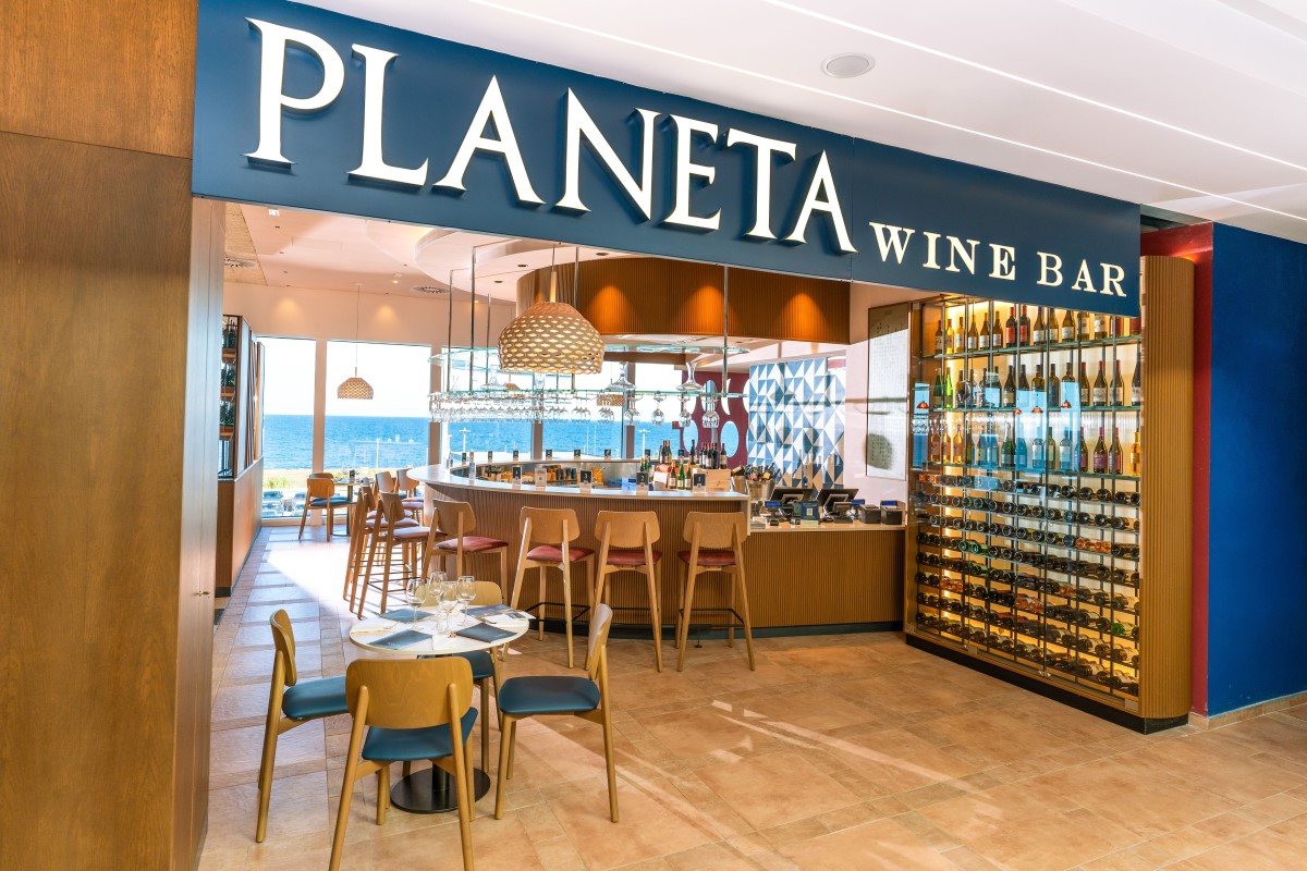 Planeta Wine Bar all'Aeroporto di Palermo: bancone, tavolini e parete di bottiglie con vista mare