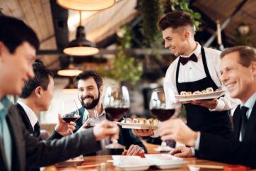 Giornata della Ristorazione: cameriere serve sushi e vino rosso a clienti sorridenti in un ristorante elegante.