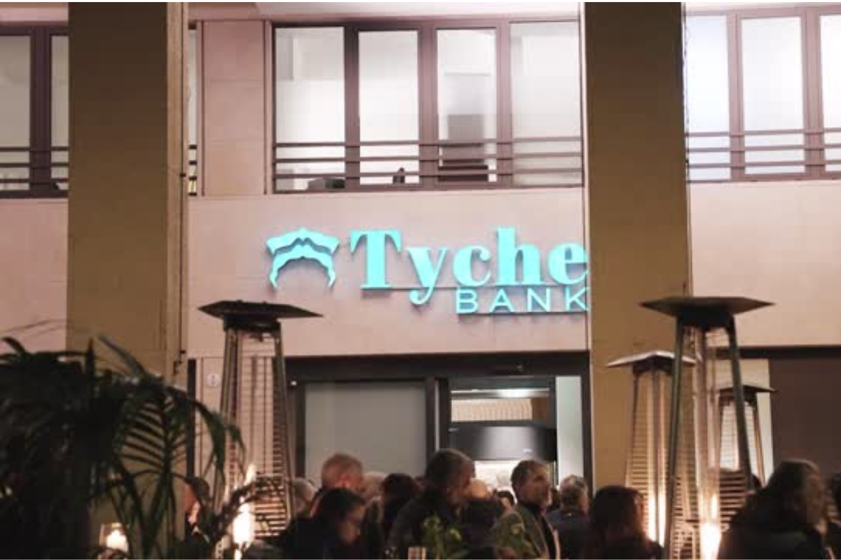 Insegna luminosa di Tyche Bank all'esterno del ristorante Madré, con persone in primo piano durante un evento serale.