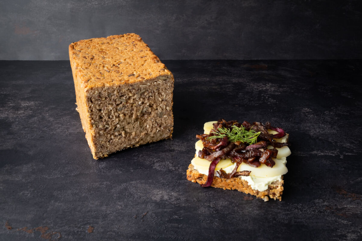 Rugbrød farcito: fetta di pane integrale ai cereali con formaggio vegano, cipolle caramellate e aneto su fondo scuro.