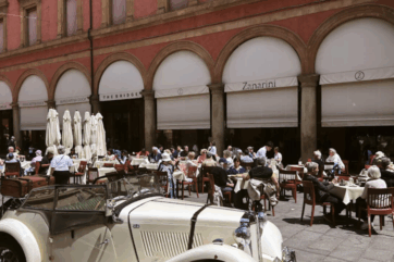 Dehors del Caffè Zanarini a Bologna con tavoli all'aperto, architettura storica e auto d'epoca in primo piano.