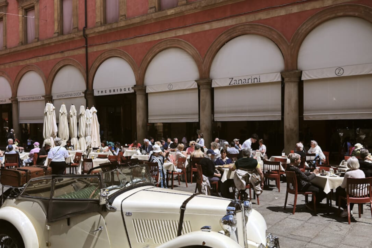 Dehors del Caffè Zanarini a Bologna con tavoli all'aperto, architettura storica e auto d'epoca in primo piano.