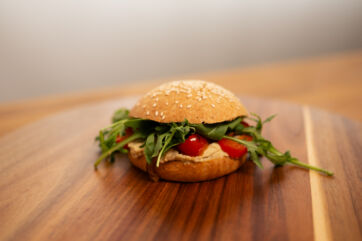 Burger bun farcito con hummus, pomodorini e rucola fresca su un tagliere in legno, ideale per una ricetta vegetariana.
