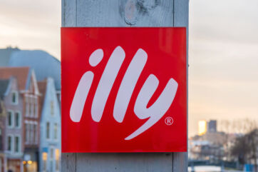 Insegna quadrata rossa con logo illycaffè su sfondo urbano, simbolo della crescita dei ricavi dell'azienda italiana.