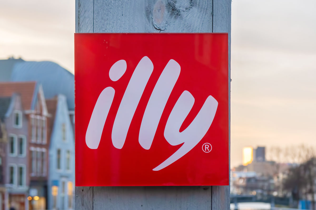 Insegna quadrata rossa con logo illycaffè su sfondo urbano, simbolo della crescita dei ricavi dell'azienda italiana.