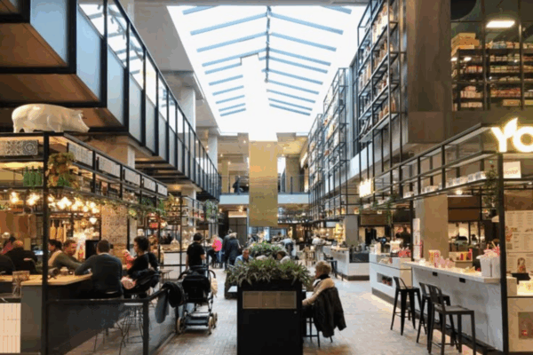 Interno moderno di una stazione ferroviaria con area food & beverage, negozi retail e clienti seduti ai tavoli.