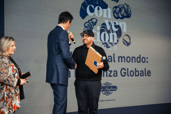 Antonio Bachour premiato sul palco dell'evento Le Sinfonie, con sfondo grafico dedicato alla pasticceria professionale.