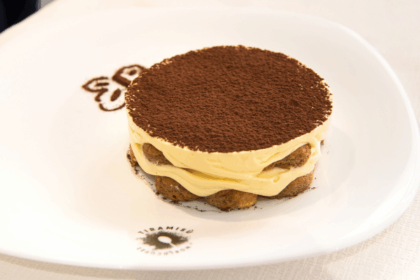 Tiramisù artigianale servito su piatto bianco con logo della Tiramisù World Cup, guarnito con abbondante cacao in polvere.