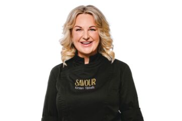 Kirsten Tibbals, la regina del cioccolato, sorridente con giacca da chef nera del brand Savour su sfondo bianco.