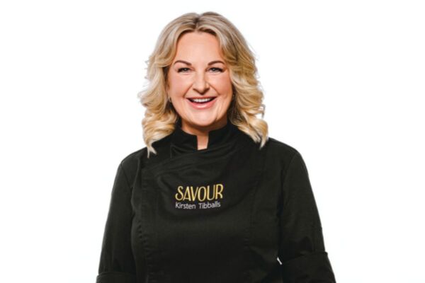 Kirsten Tibbals, la regina del cioccolato, sorridente con giacca da chef nera del brand Savour su sfondo bianco.