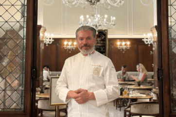 Domenico Di Clemente, chef in divisa bianca, posa all'interno di un elegante ristorante di lusso con lampadari classici.
