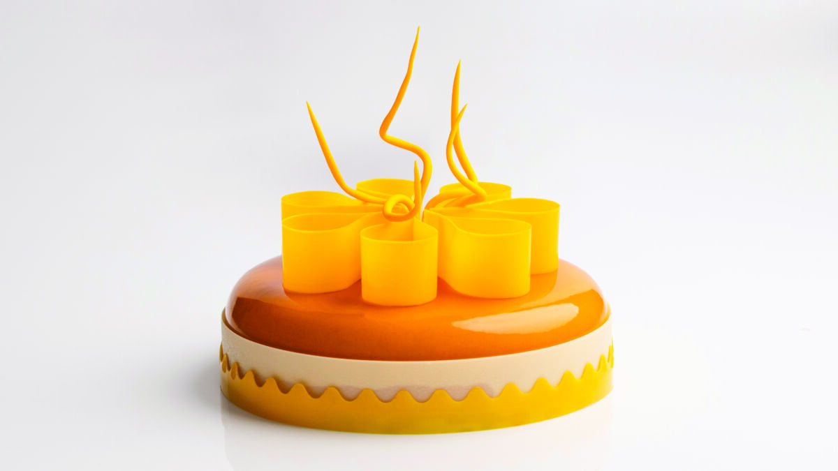Torta moderna Fleur con glassa arancione lucida, decorazioni in cioccolato giallo a forma di fiore e base smerlata.