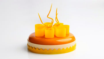 Torta moderna Fleur con glassa arancione lucida, decorazioni in cioccolato giallo a forma di fiore e base smerlata.