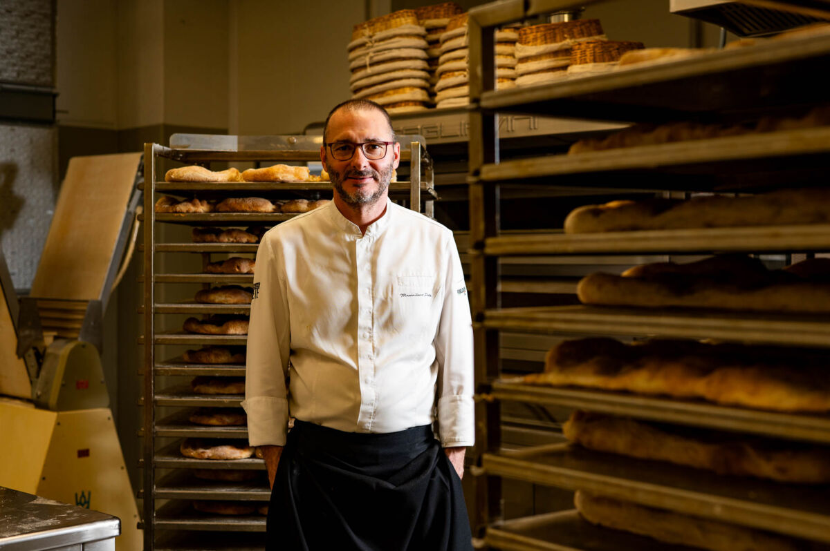 Massimiliano Prete, chef panificatore, posa sorridente tra scaffali di pane appena sfornato nel suo laboratorio.