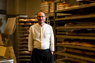 Massimiliano Prete, chef panificatore, posa sorridente tra scaffali di pane appena sfornato nel suo laboratorio.