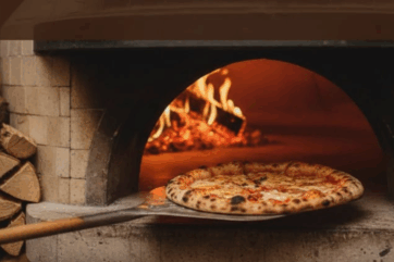 Pizza appena sfornata su pala in legno all'interno di un forno a legna per la finale della Pizza Bit Competition 2026.