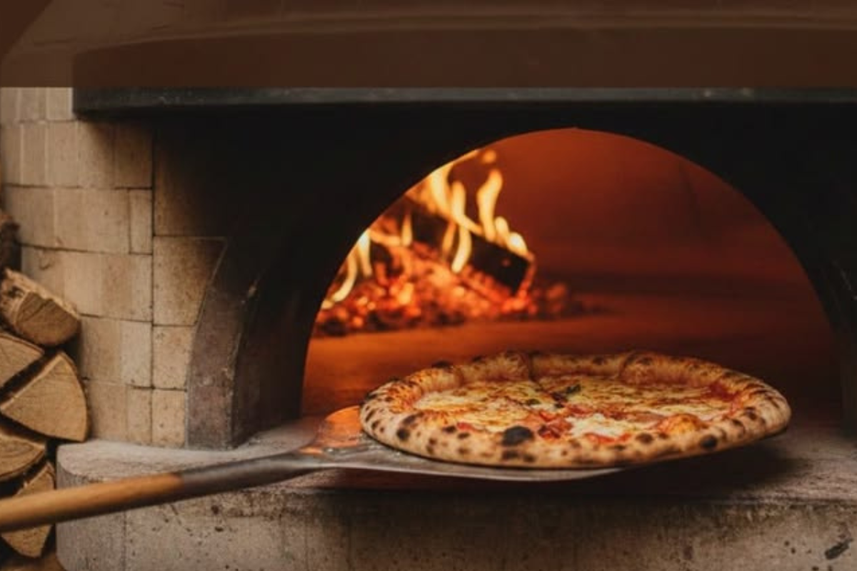 Pizza appena sfornata su pala in legno all'interno di un forno a legna per la finale della Pizza Bit Competition 2026.