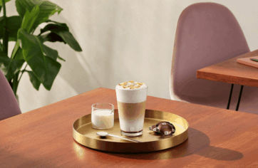 Caffè nel fuori casa: latte macchiato cremoso su vassoio dorato con tavolo in legno e sedia rosa, esperienza di gusto