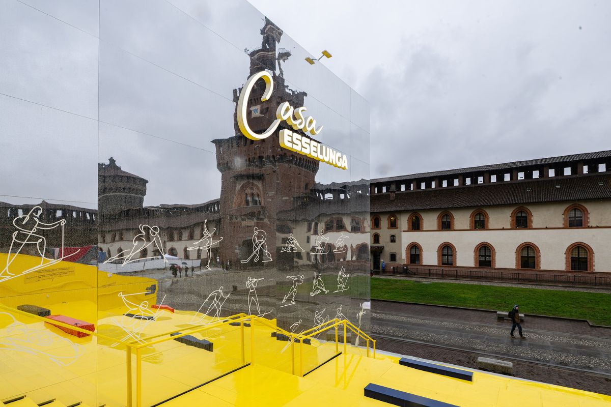Casa Esselunga Milano Cortina 2026: riflesso del Castello Sforzesco su parete con scritte e disegni sportivi.