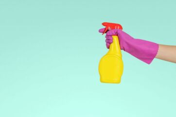 Detersivo spray: mano con guanto viola tiene flacone giallo. Pulizia sicura, stop test animali.