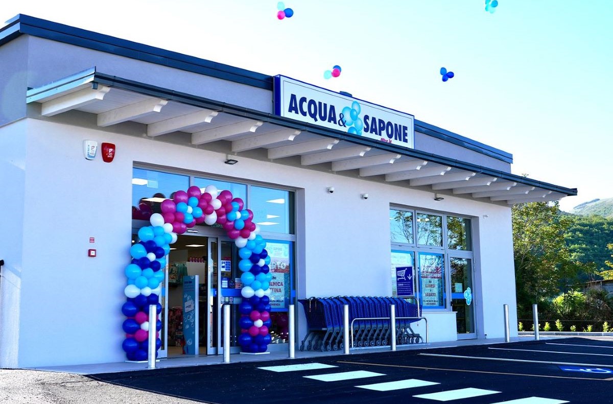 Negozio Acqua & Sapone. Facciata bianca con arco di palloncini colorati all'ingresso. Carrelli spesa in fila.