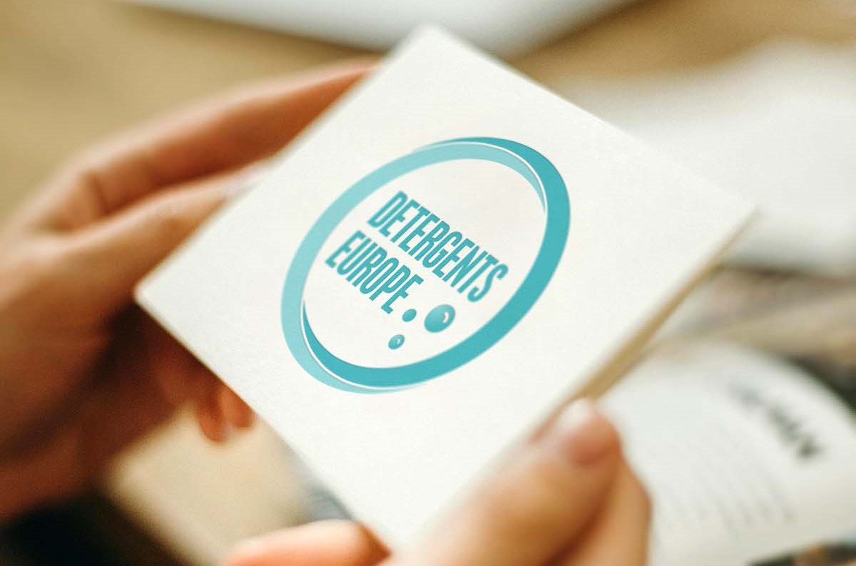 Mani che tengono un cartoncino bianco con il logo blu di Detergents Europe, nuovo nome dell'associazione A.I.S.E.
