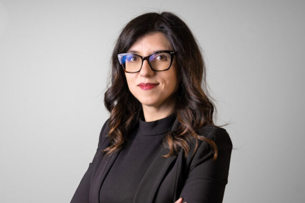 Ritratto professionale di una manager di Bolton Home Care con giacca nera e occhiali su sfondo grigio neutro.