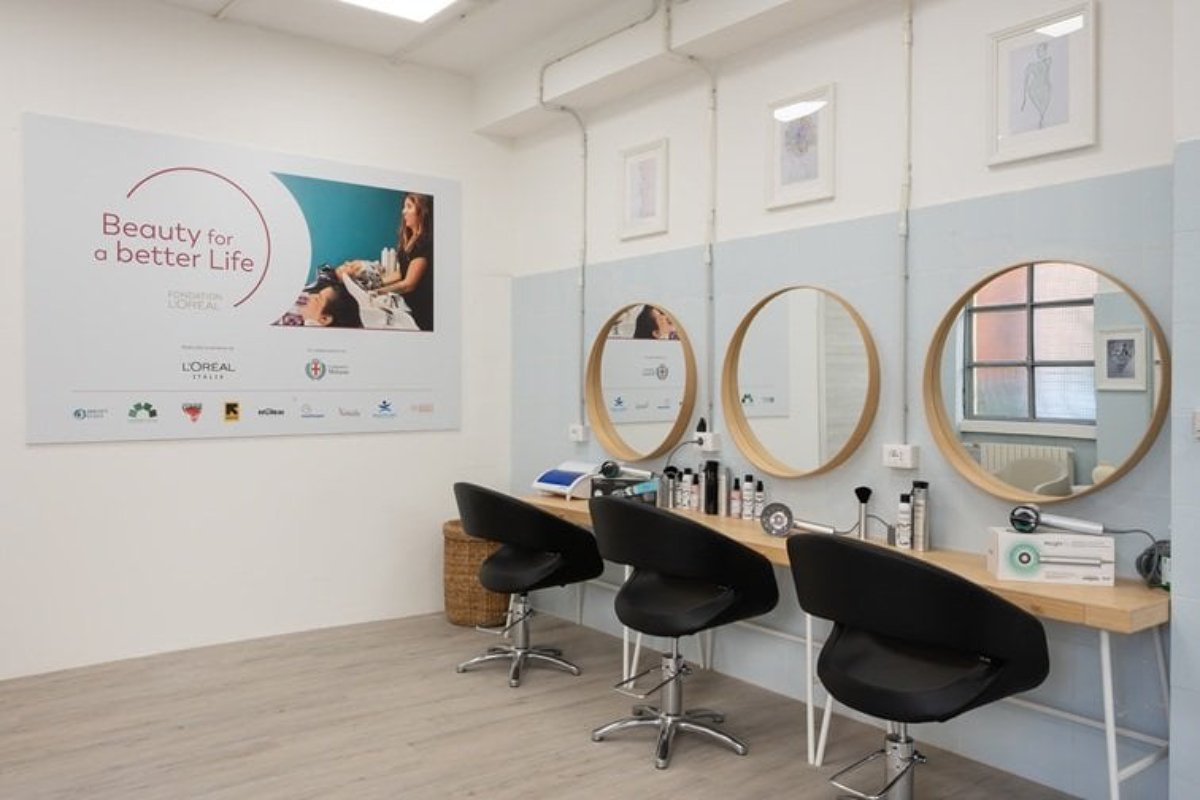 Salone sociale L'Oréal: postazioni parrucchiere con specchi rotondi e sedie nere. Poster Beauty for a Better Life.