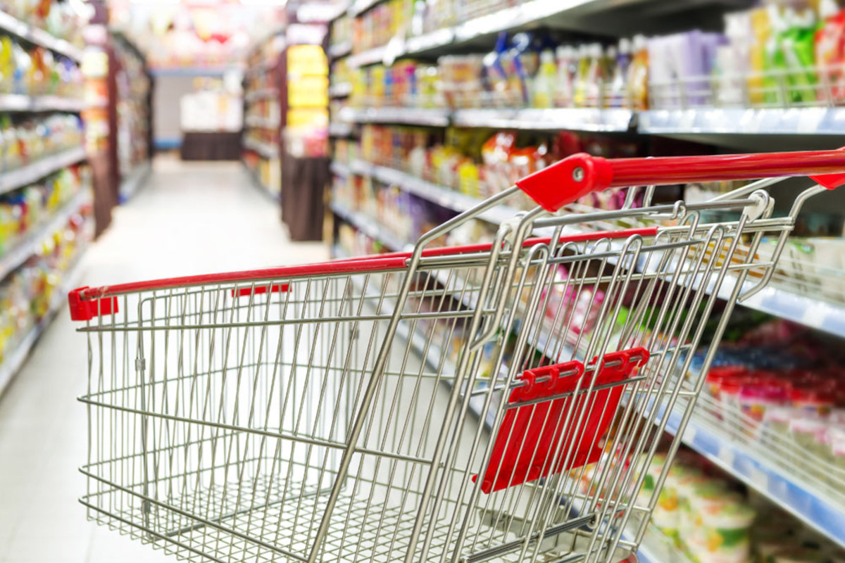 Carrello spesa rosso vuoto in supermercato. Corsie con scaffali pieni di prodotti alimentari.