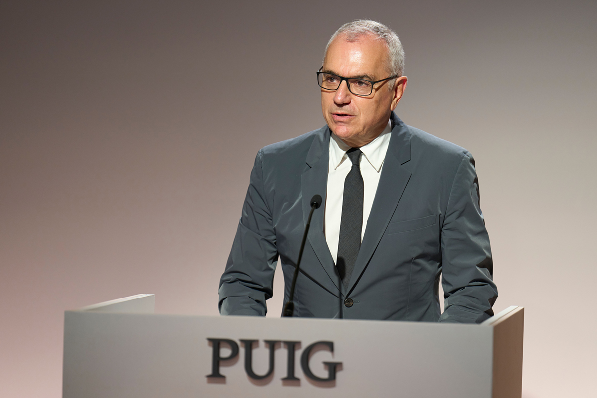 Marc Puig, presidente del Gruppo Puig, parla a un evento aziendale dietro un podio con il logo del brand in evidenza.